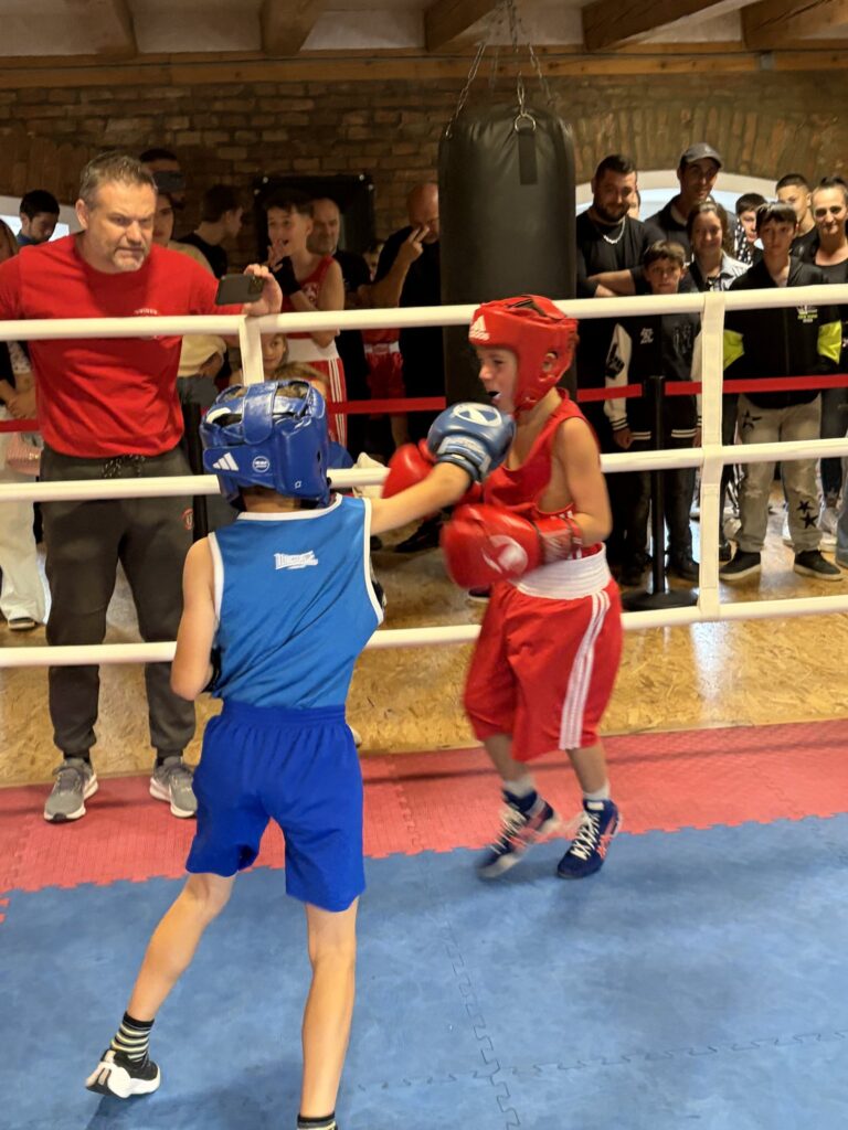 Mladí boxeri v modrom a červenom počas zápasu, pri ringu tréner